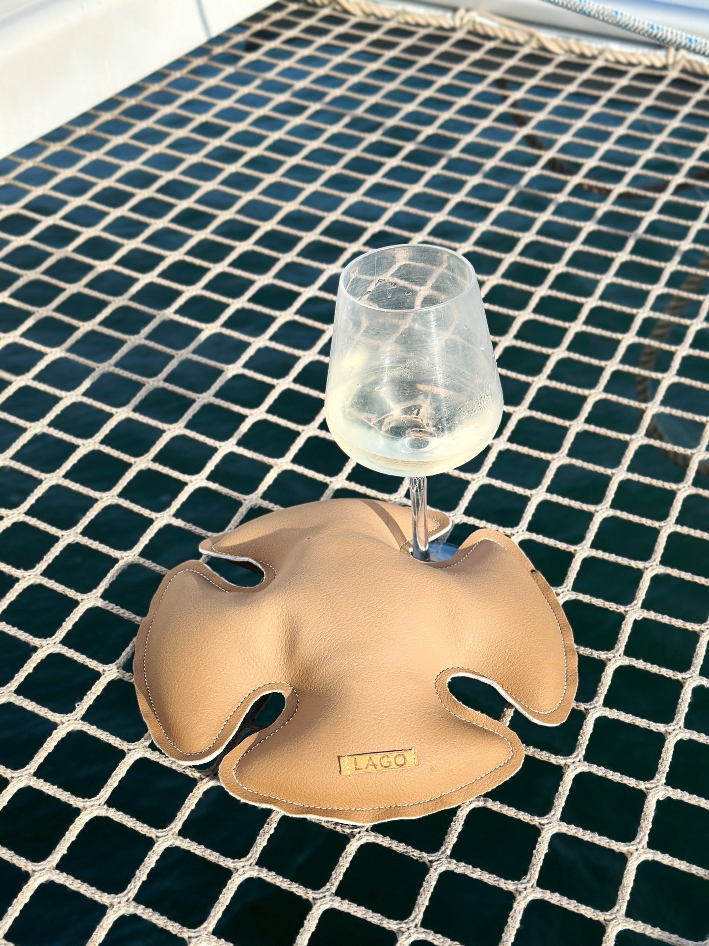 LAGO wine glass stabilizer in desert tan marine vinyl, elegantly securing a stemmed glass on a boat’s netted deck with clear blue water beneath.