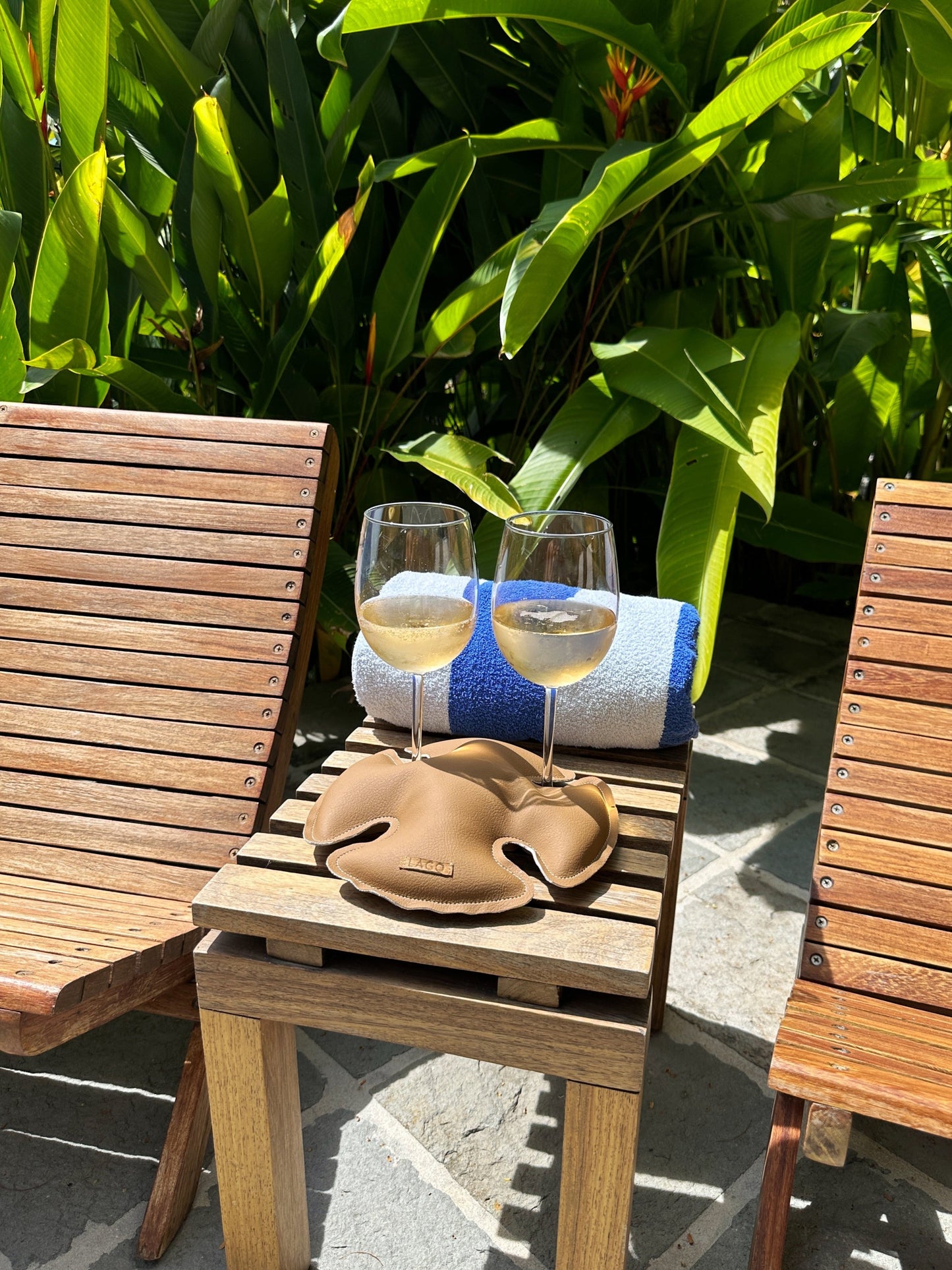 Two glasses of chilled wine poised on a LAGO stabilizer between wooden chairs, ready for a relaxing poolside retreat amidst lush tropical foliage.